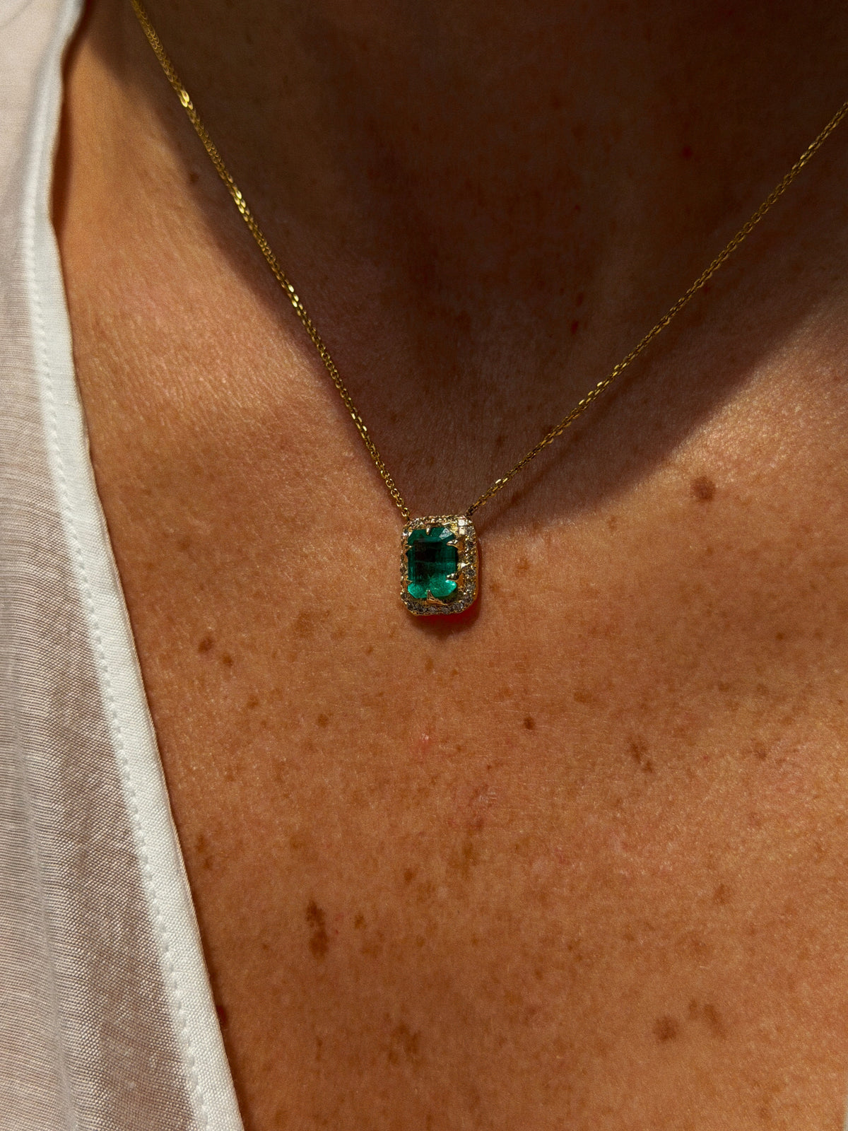 North-South emerald-cut green emerald necklace with diamond halo and 14K yellow gold chain, worn at the neckline.