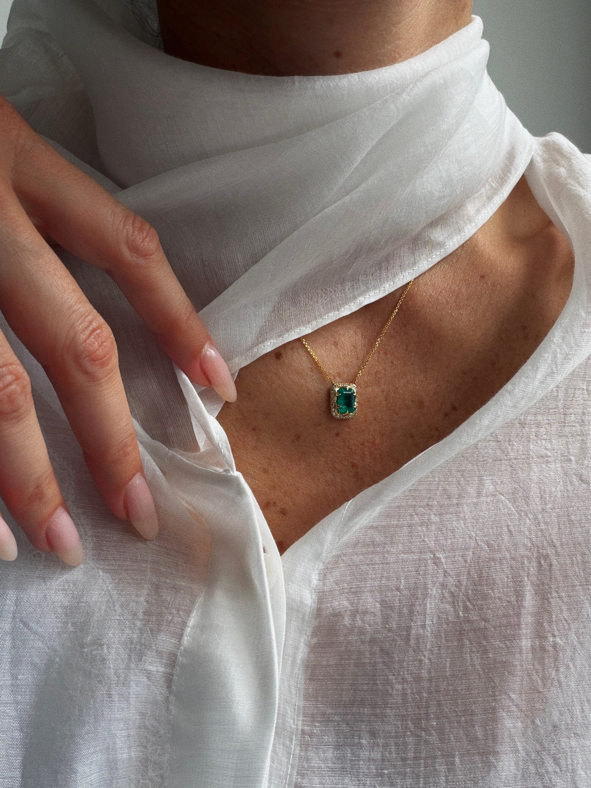 Close-up of a green emerald-cut emerald and diamond halo necklace in 14K yellow gold, worn with a white blouse and scarf.