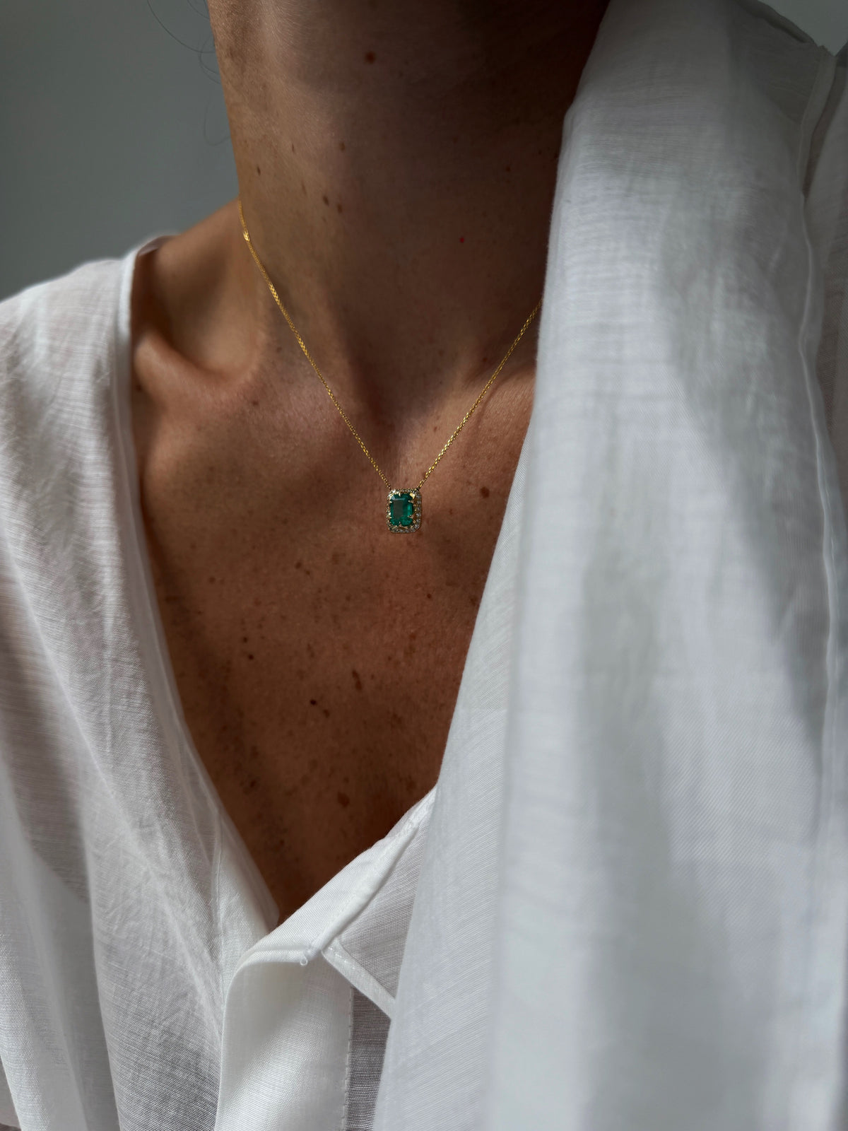 Emerald-cut emerald pendant necklace with diamond halo in 14K yellow gold, styled with a white blouse.