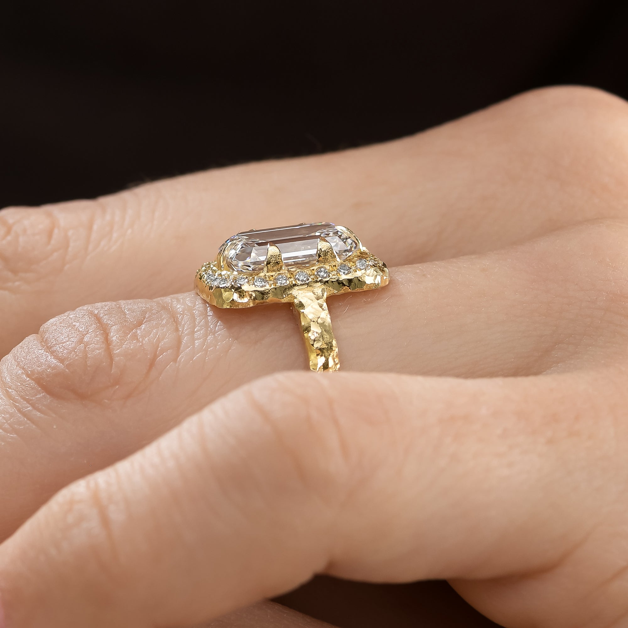 Angled view of 4 ct emerald-cut diamond engagement ring with textured 14K yellow gold setting and diamond halo worn on hand.