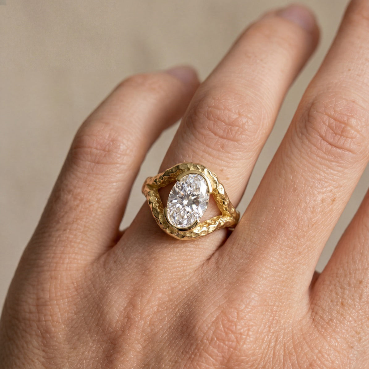 A close-up of a hand wearing a unique gold ring with a large oval clear gemstone in the center, set against a soft beige background.