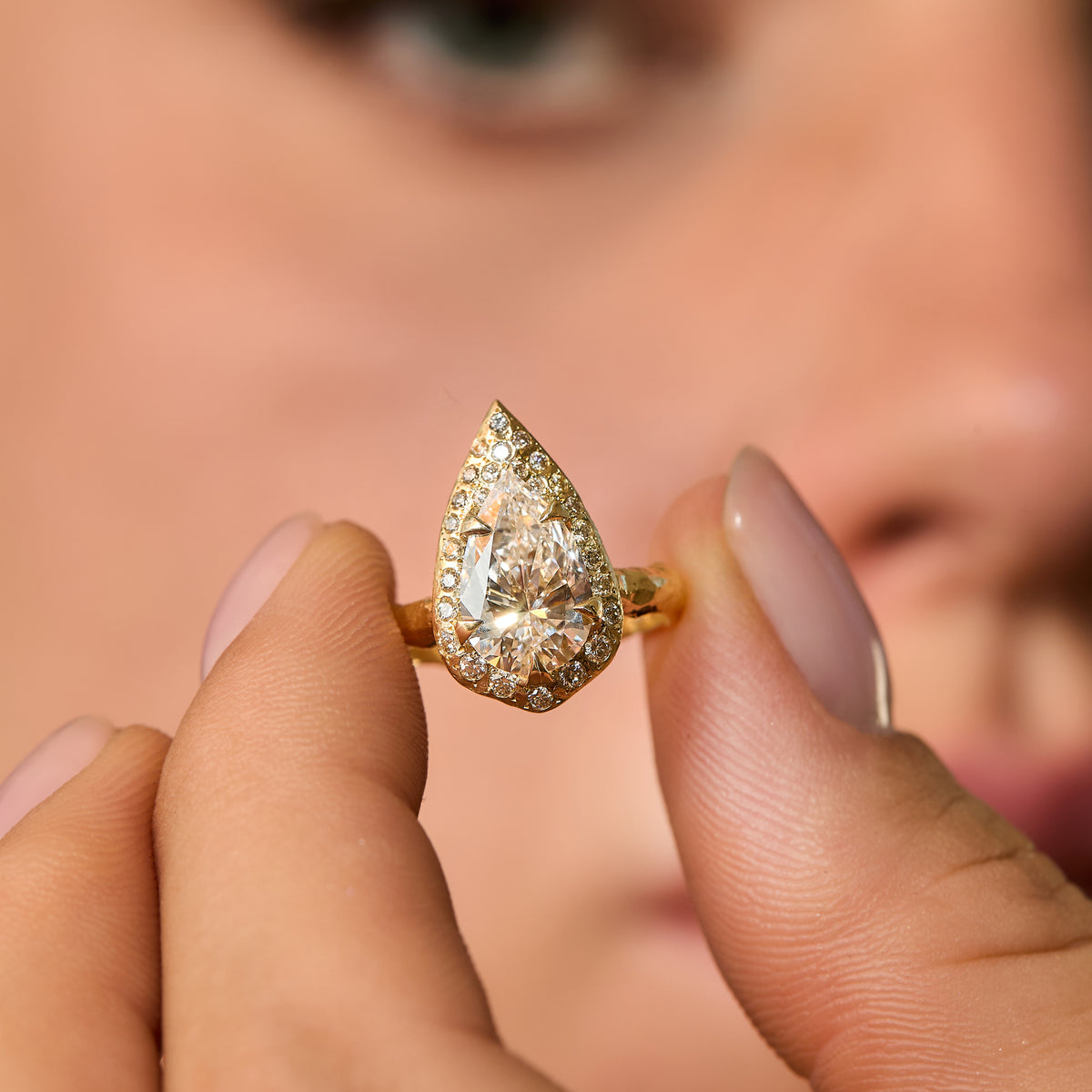ring with 2 CT pear-shaped natural diamond engagement with diamond halo and hand-carved organic gold band hold between fingers.