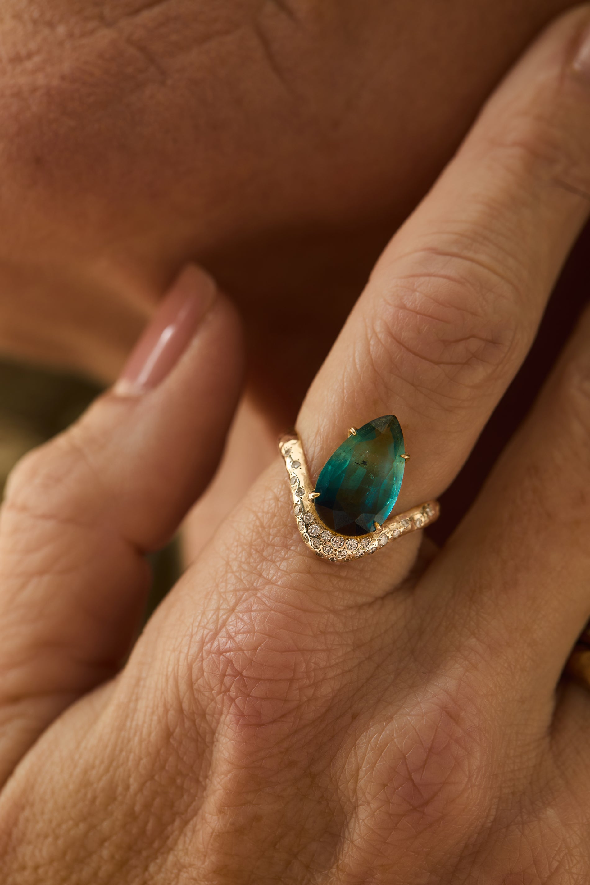 A close-up of a hand wearing a gold ring with a pear-cut teal-green multicolor gemstone and small diamondson the band